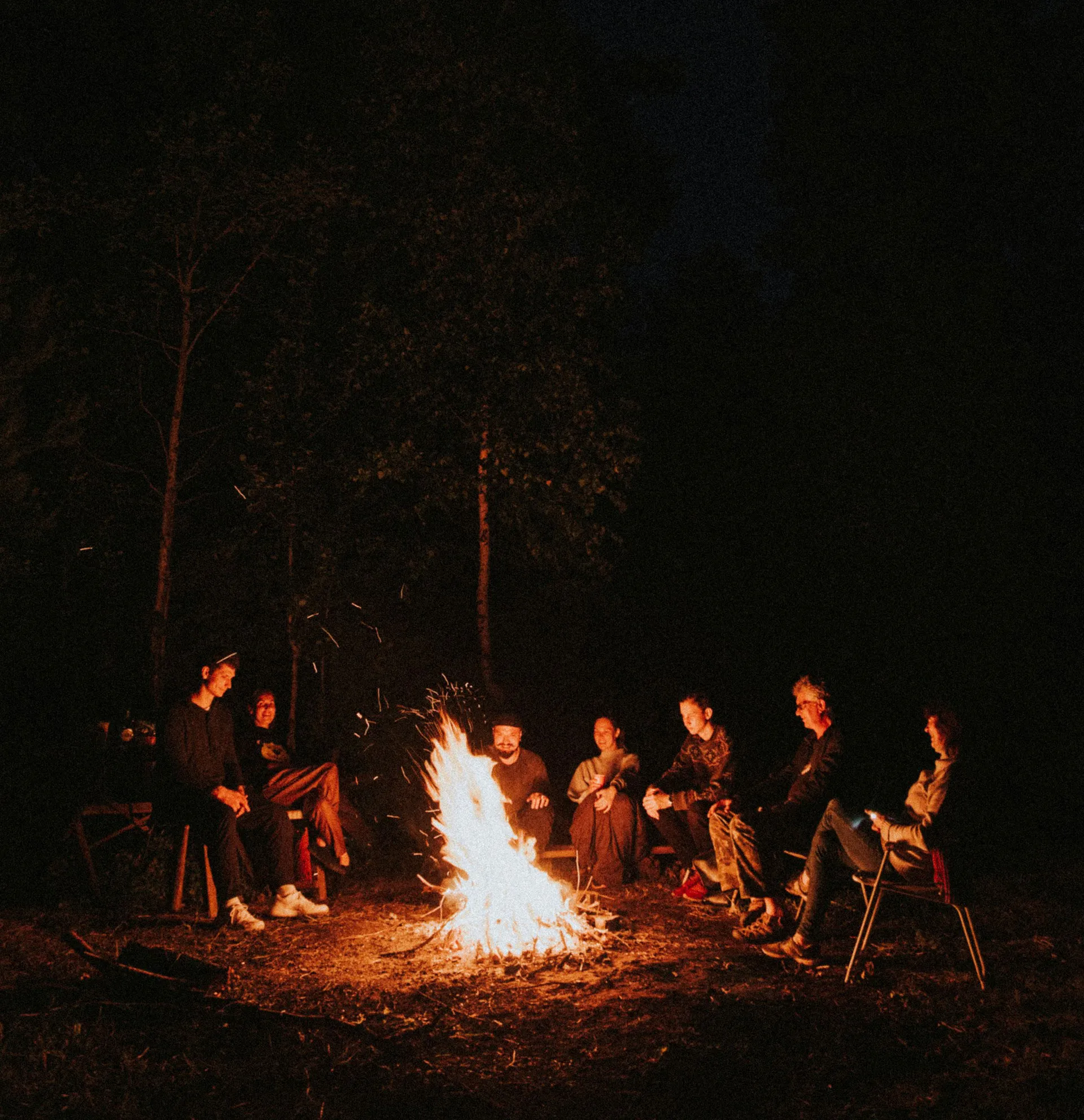 Warm community gathering around a crackling fire at dusk, showing people connecting and enjoying each other's company in a cozy outdoor setting